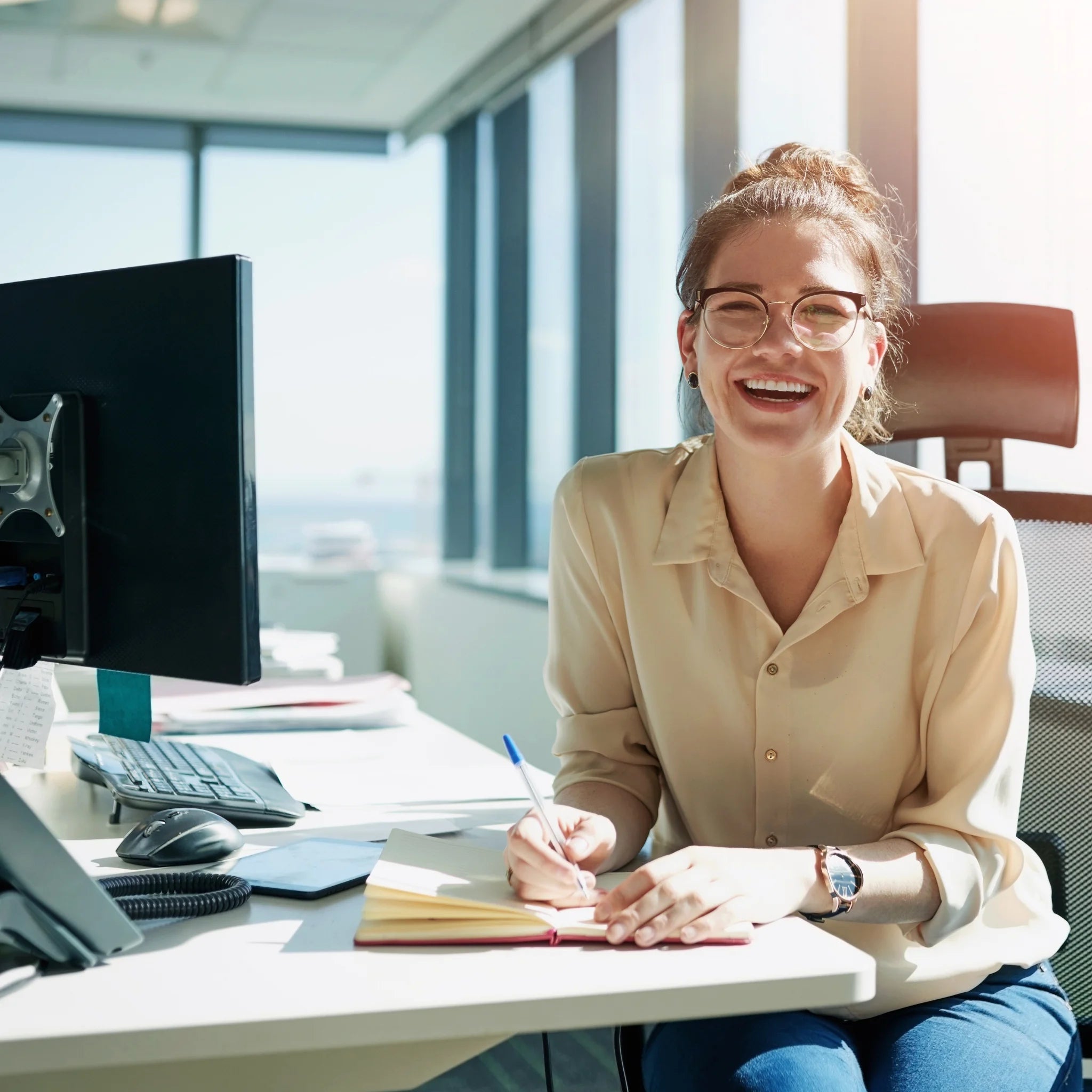 Junge, lächelnde Frau mit Brille und Dutt sitzt an ihrem Büroschreibtisch an einem sonnigen Fenster und schreibt Notizen in ein rotes Notizbuch. Sie trägt eine beige Bluse und blickt optimistisch in die Kamera.