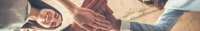 Aufsicht auf sechs lachende Gesichter einer vielfältigen Gruppe von Menschen, die ihre Hände in der Mitte stapeln. Symbolisiert Teamwork, Einheit und Zusammenhalt nach einem Projektabschluss.