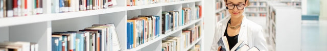 Eine junge Frau mit Brille und einem weißen Anzug steht in einer hellen Bibliothek und hält mehrere Bücher. Das Bild zeigt lange Reihen weißer Bücherregale, die mit vielen Büchern gefüllt sind.