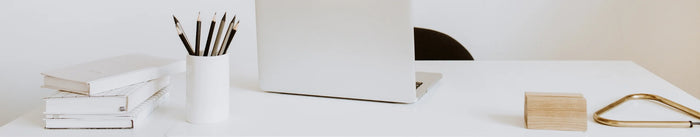 Ein minimalistischer, heller Schreibtisch mit einem silbernen Laptop, einem Stiftehalter mit Bleistiften und einem Stapel weißer Skillbooks (Notizbücher).