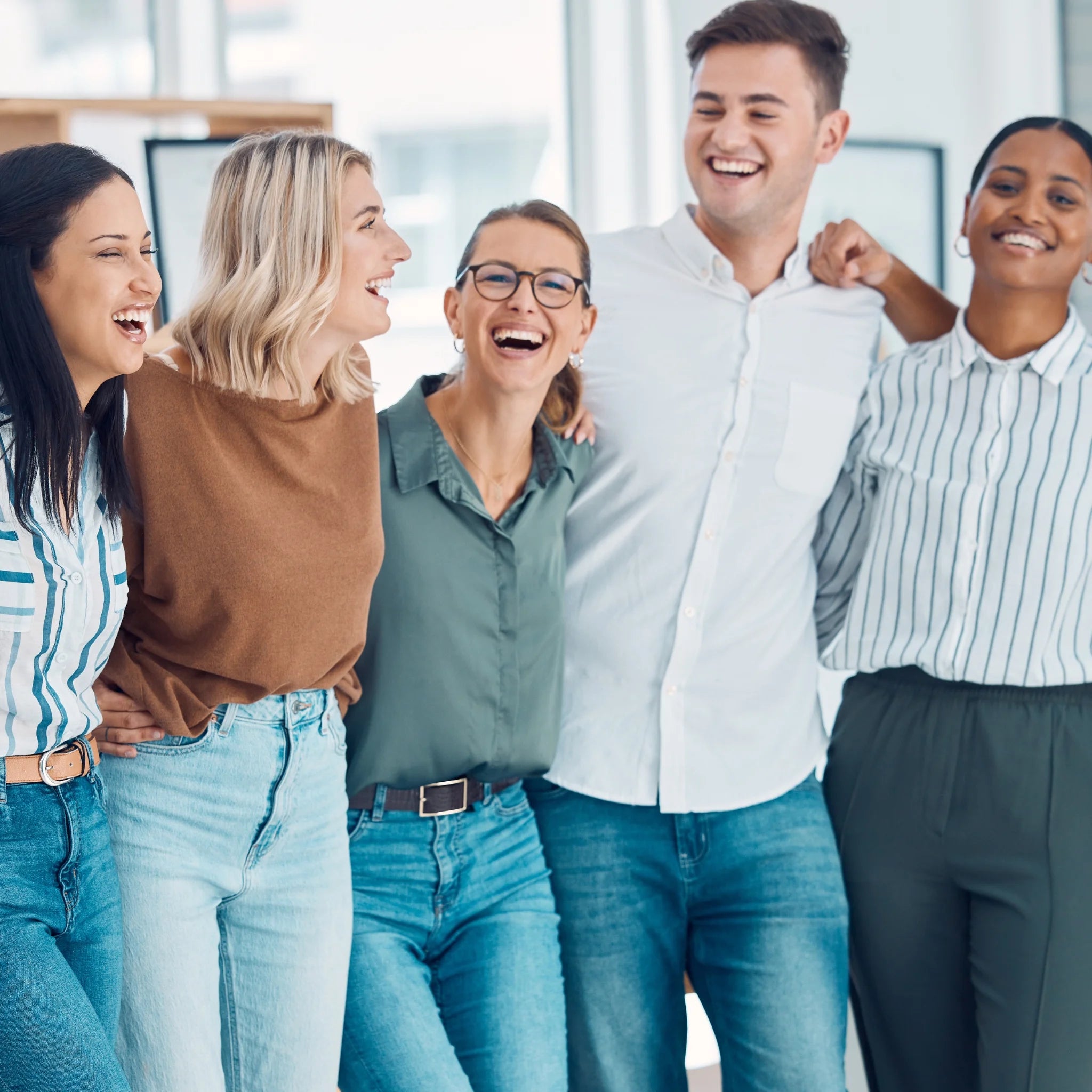 Eine vielfältige Gruppe von fünf jungen, lachenden Mitarbeitern steht eng beieinander und umarmt sich gegenseitig im Büro. Zeigt positives und harmonisches Teamwork.