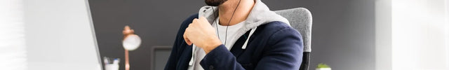 Ein lächelnder Mann mit Brille und Headset sitzt an einem weißen Schreibtisch vor seinem Computer und Laptop. Er trägt einen Kapuzenpullover unter einem Sakko und symbolisiert aktiven Online-Workshop.