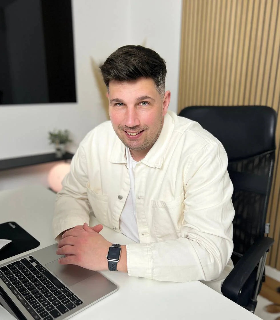 Ein lächelnder Mann, Sebastian, in einer weißen Jacke sitzt an einem hellen Schreibtisch vor seinem Laptop. Er blickt direkt in die Kamera und vermittelt den Eindruck eines Beraters oder Coaches. 