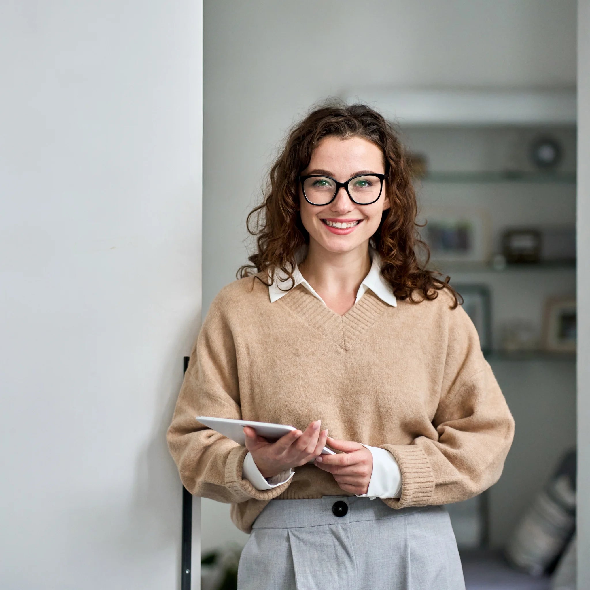 Junge, lächelnde Frau mit lockigen Haaren und Brille, hält ein Tablet und trägt einen beigefarbenen Pullover und eine graue Hose.
