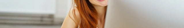 Eine strahlend lächelnde Frau mit langen, roten Haaren blickt hinter einem silbernen Computermonitor hervor. Sie trägt ein helles Oberteil und symbolisiert freundlichen Kunden- oder Online-Service.