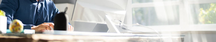 Nahaufnahme der Hände eines Mannes in einem blauen Hemd, der an einem modernen Computer arbeitet. Die Szene spielt in einem hellen Büro am Fenster mit viel Sonnenlicht und suggeriert digitales Arbeiten und Produktivität.