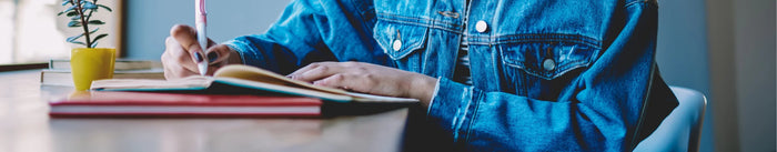 Nahaufnahme einer Person in einer Denim-Jacke, die mit einem Stift in einem offenen Skillbook (Notizbuch) schreibt. Mehrere rote und braune Bücher liegen auf dem Schreibtisch.